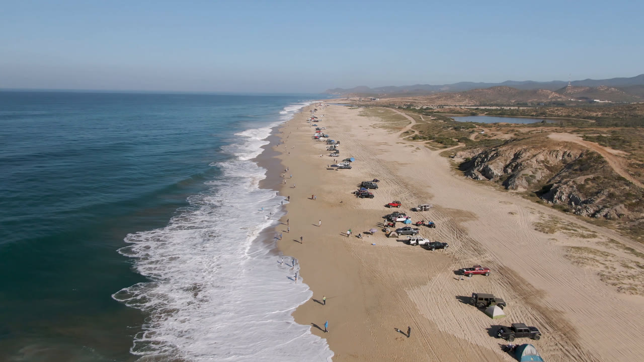 Drone view along a sandy coast with surf, hills, and clusters of cars on the beach.