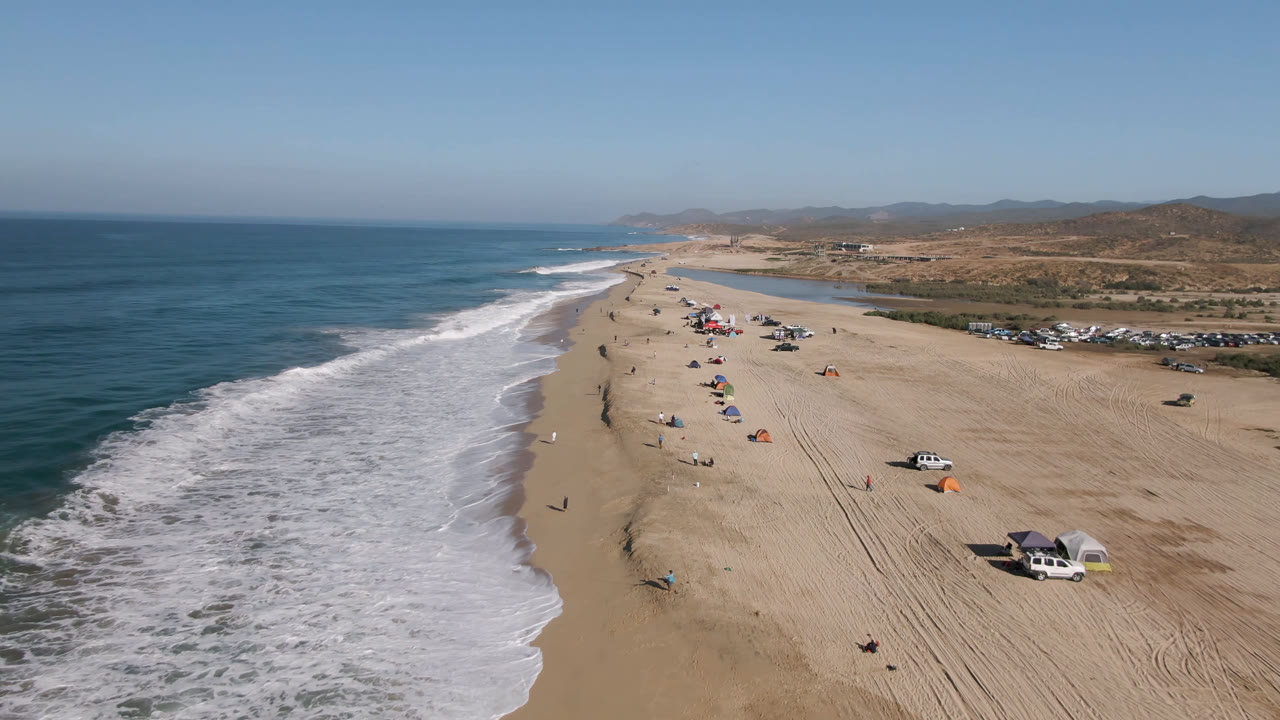 Aerial view over Arafly Beach in Migrino, Baja California Sur, with white surf at left, wide sand, scattered umbrellas, parked vehicles, and low inland hills in clear daylight.
