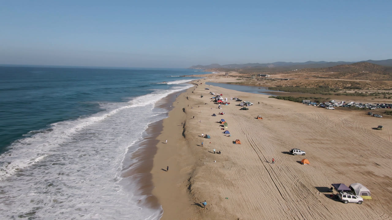 Drone view along Arafly Beach in Migrino, Baja California Sur, showing the surf line curving into the distance, a central strip of beachgoers and shade umbrellas, and vehicles parked near the dunes.