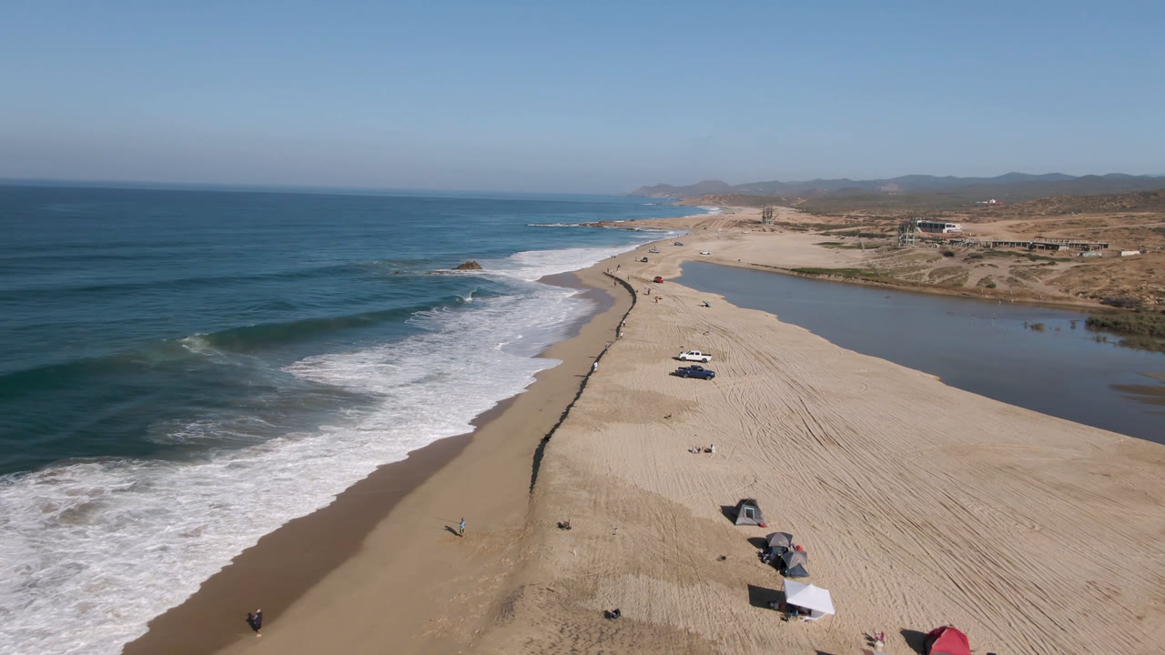 Aerial view over Migrino, Baja California Sur with blue ocean surf on the left, a sandy beach with vehicles and small people below, and a lagoon running along the right side.