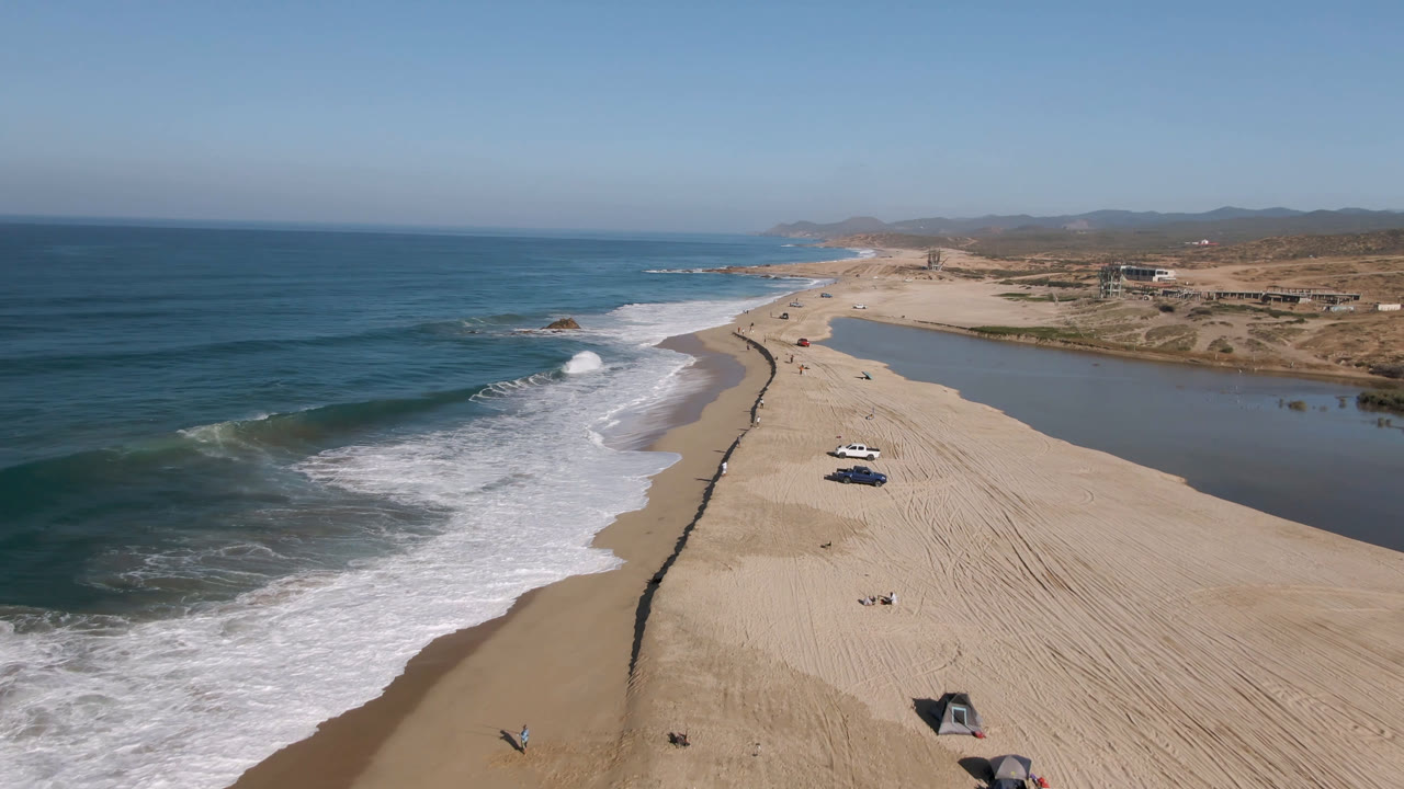 Migrino beach from above with a fresh wave line curling toward the sand, parked vehicles near the lower beach, small people on the shoreline, and a long lagoon beside the dunes.