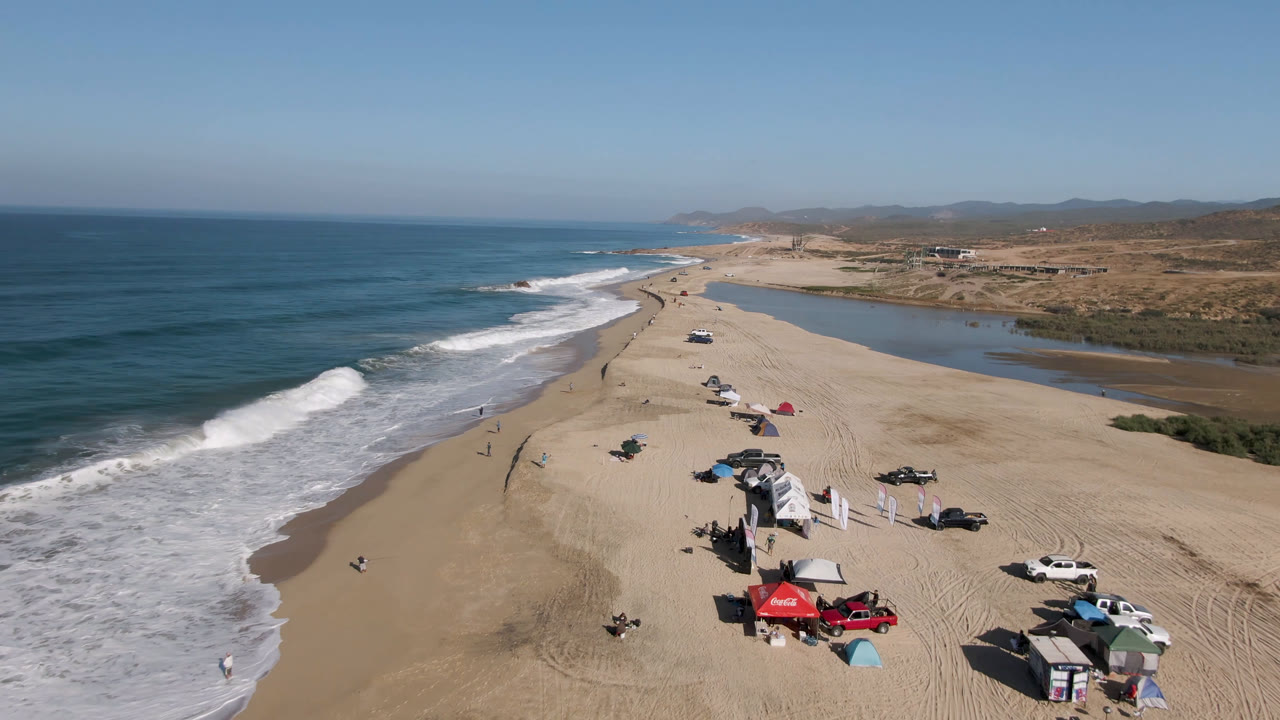 Aerial frame from San Jose Del Cabo, Mexico showing breaking surf on the left, a sandy camping area with vehicles and tents below, and lagoon water behind the beach.