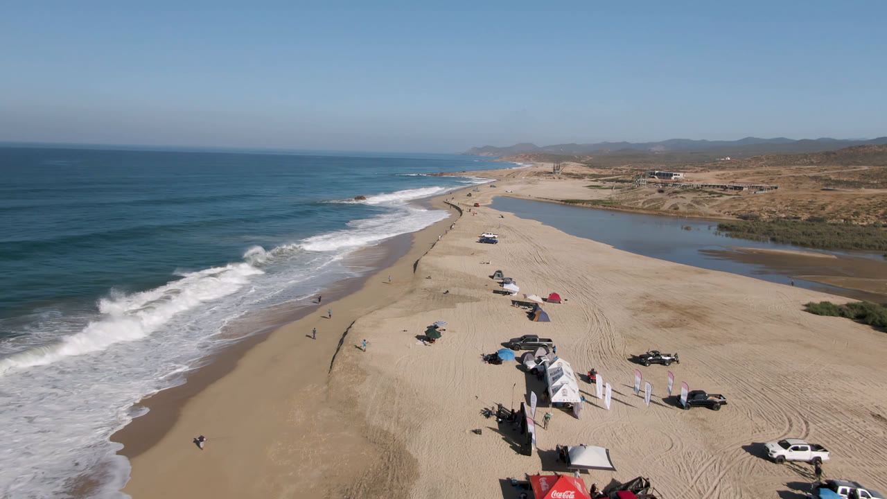 Wide aerial view in San Jose Del Cabo, Mexico with white surf advancing along the shore, campsites and vehicles on the sand, and a lagoon running parallel to the beach.
