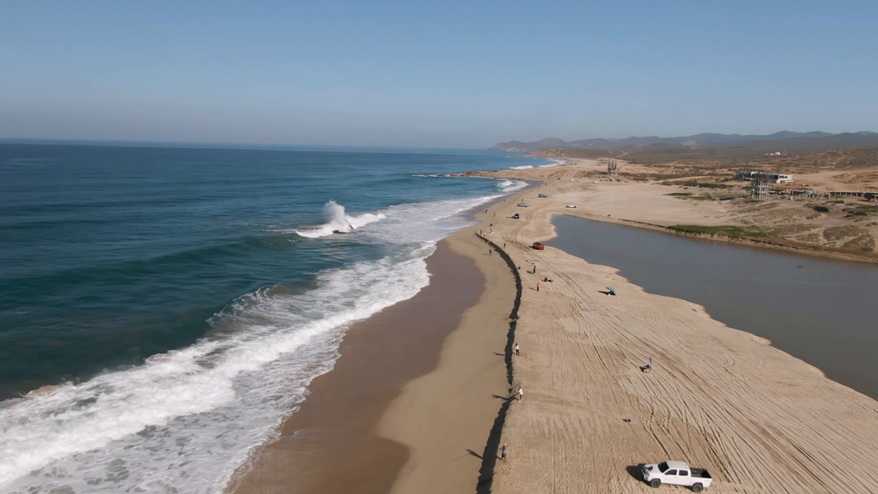 Aerial frame over Migrino, Baja California Sur, with blue ocean and breaking surf on the left, a sandy beach and dark shoreline line through the middle, lagoon water on the right, and small distant people visible.