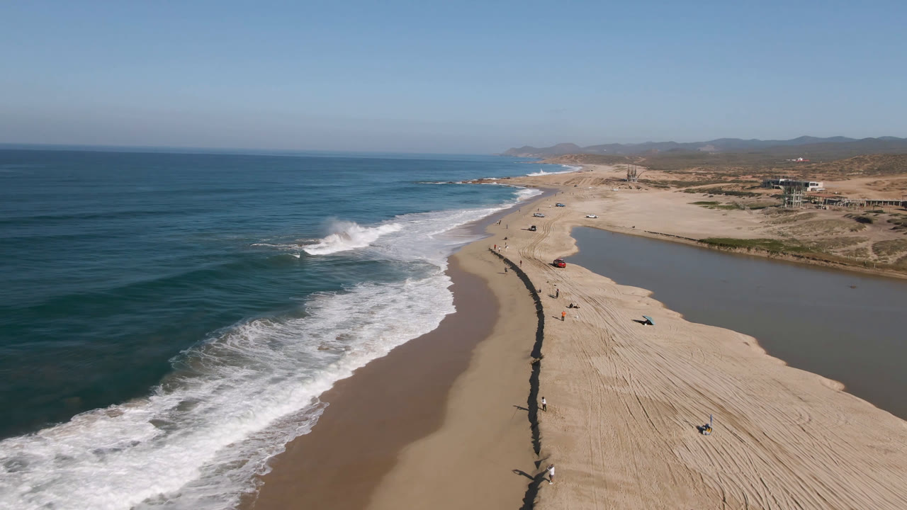 Aerial view of Migrino beach with the surf line closer to center, pale sand stretching toward distant hills, lagoon water on the right, scattered vehicles, and tiny people visible along the dark beach line.
