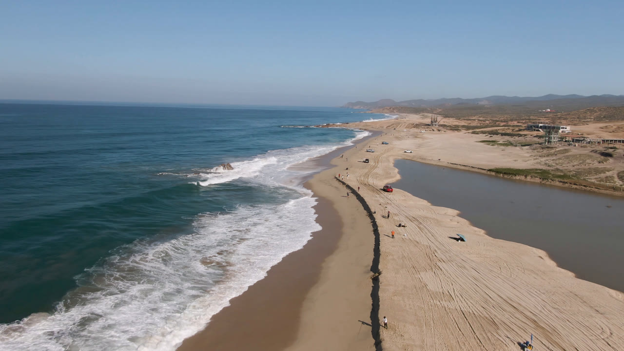 Wide aerial beach view with waves, sand, lagoon water, and distant hills.