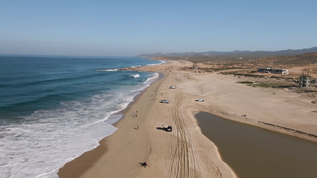 Aerial view of ocean surf, sandy beach, vehicle tracks, parked cars, and a lagoon edge.