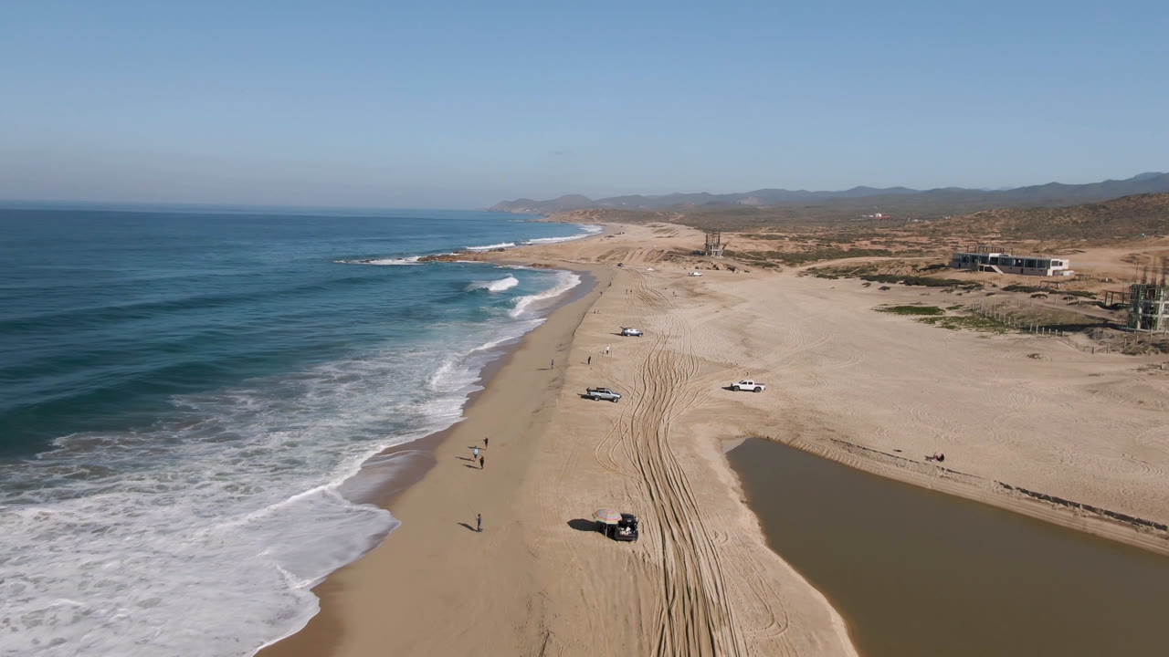 Aerial frame from Migrino, Baja California Sur, showing the surf line closer to center, a long sandy beach with parallel tire marks, scattered vehicles, low scrub, and hills beyond the coast.