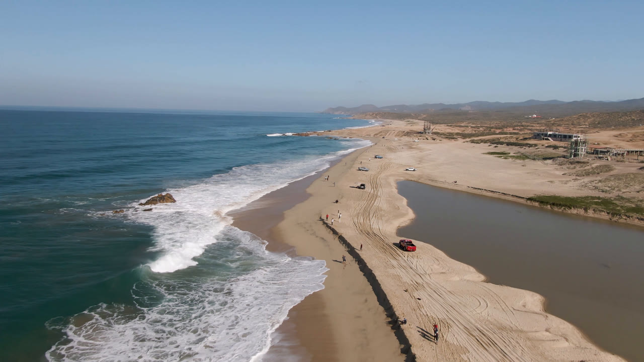 Aerial frame over Migrino, Baja California Sur, Mexico showing ocean surf on the left, a sandy beach with tire tracks, a red truck near the lower right, and a lagoon along the inland side.