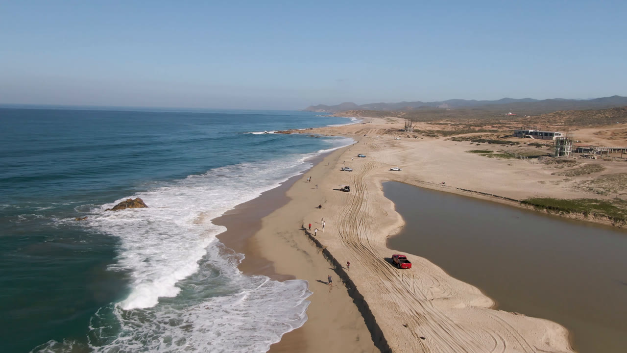 Aerial view along the Migrino shoreline with the red truck farther up the sandy beach, white surf rolling in from the left, vehicle tracks in the sand, and low hills under clear daylight.