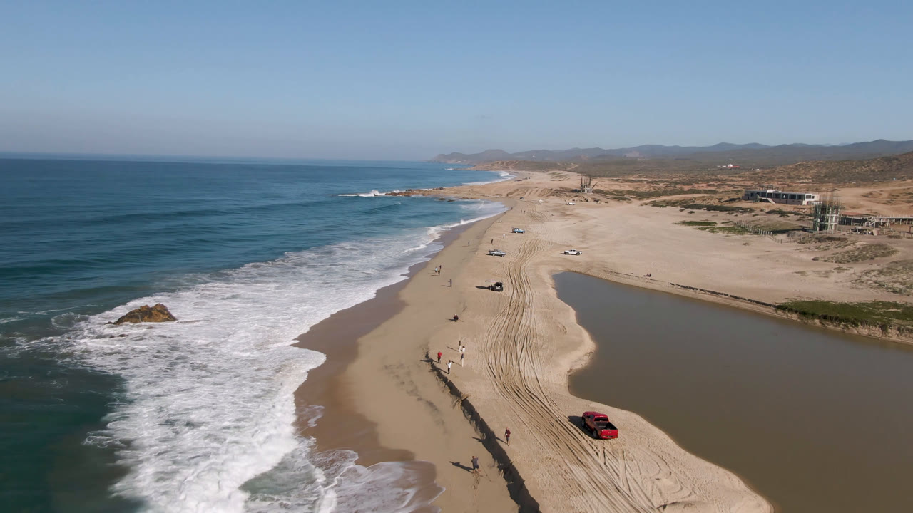 Drone view of waves, beach tracks, a red truck, and open coastline.