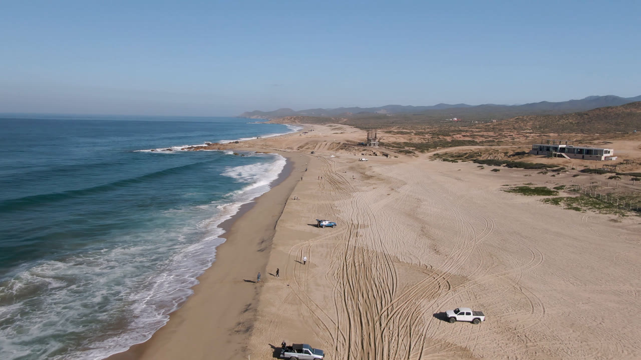 Aerial view in Migrino, Baja California Sur, Mexico, with a blue truck on open sand near the surf, tire tracks across the beach, coastal hills in the background, and no visible people.