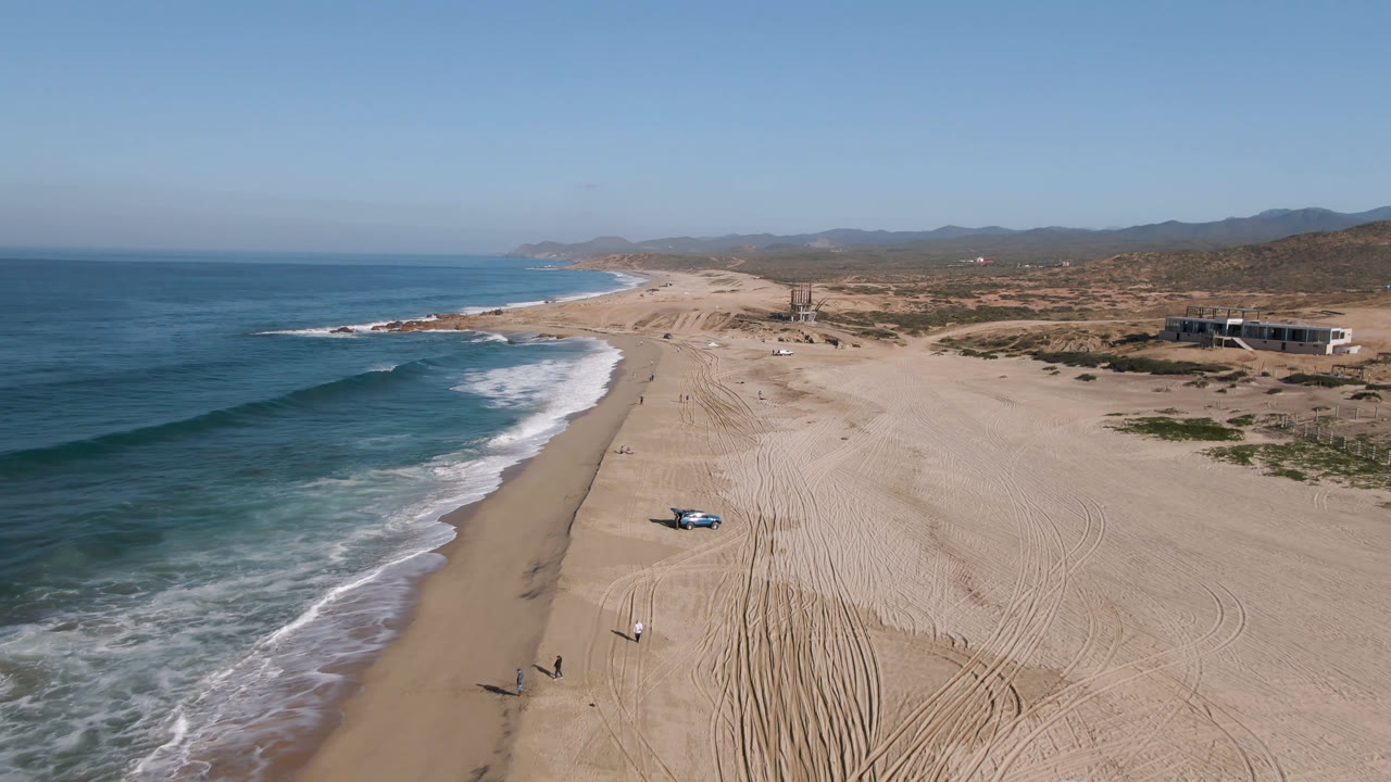 Drone-height view over Migrino beach showing the blue truck closer to the lower center of the frame, breaking waves on the left, pale sand tracks, distant hills, and no visible people.