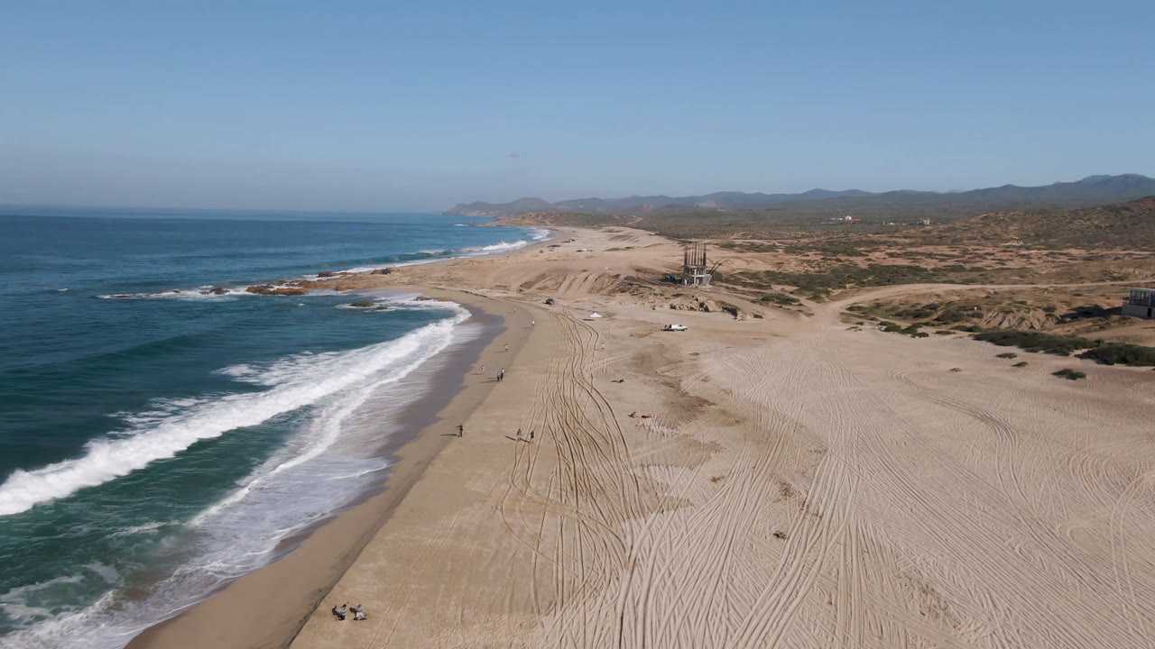 Aerial view in Migrino, Baja California Sur with the surf edge closer to the lower left, tire-marked sand running toward the horizon, dry inland terrain, and no visible people.