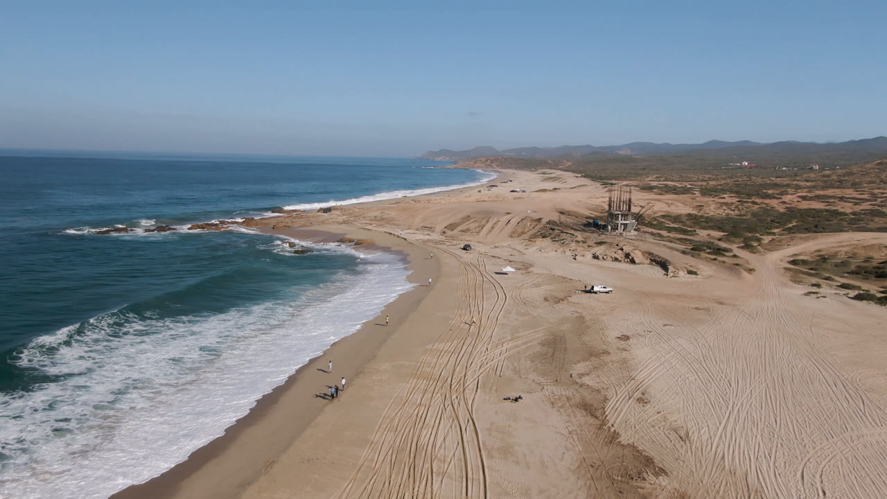 Aerial frame over Migrino, Baja California Sur, showing blue surf on the left, broad sandy beach with tire tracks, a small truck near the shoreline, dunes beyond, and no visible people.