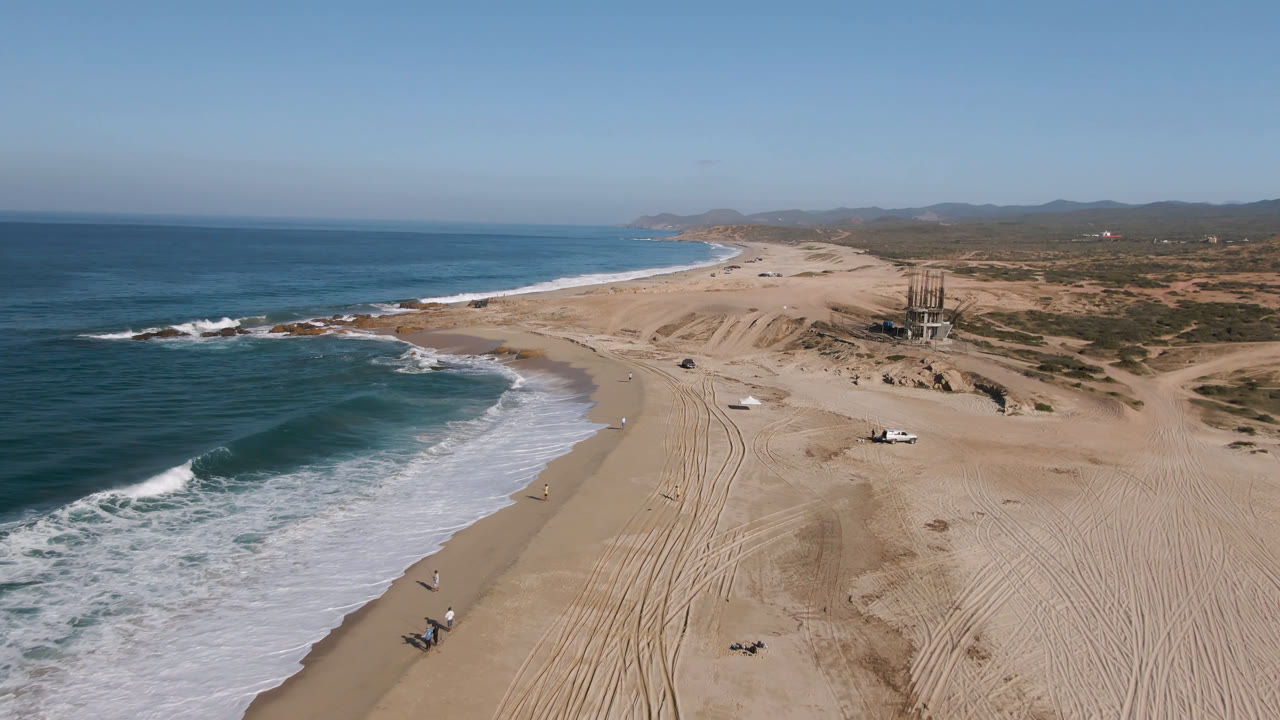 Drone view over Migrino beach with the foamy wave edge lower left, tire tracks running up the sand, a small truck farther inland, dunes to the right, and no visible people.