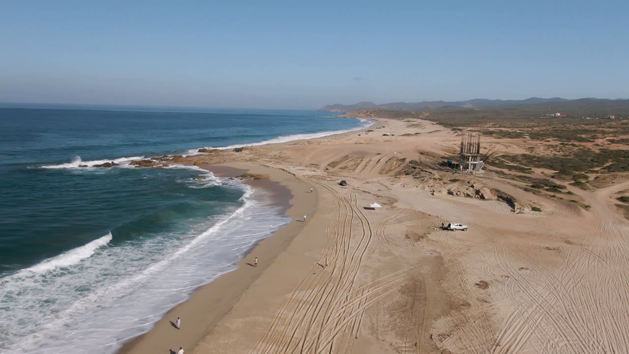 Aerial coastline with waves, beach tracks, dunes, and distant structures.