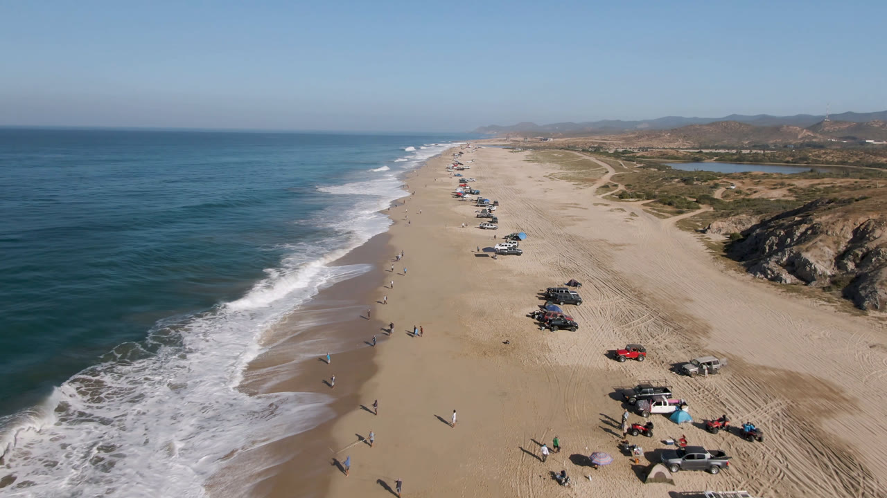 Aerial beach view with ocean surf, parked cars, and dunes