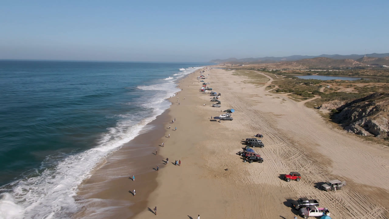 A second aerial angle over Arafly Beach shows the surf line stretching into the distance, rows of vehicles on the sand, small distant people near the water, and scrubby dunes inland.