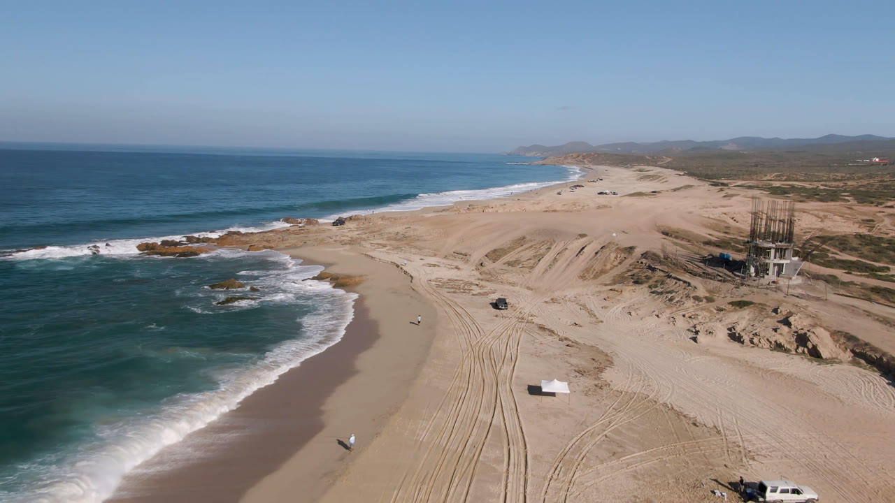 Aerial frame over Migrino, Baja California Sur, Mexico, showing ocean surf on the left, wide sand with tire tracks, a parked truck near the lower edge, distant hills, and no visible people.