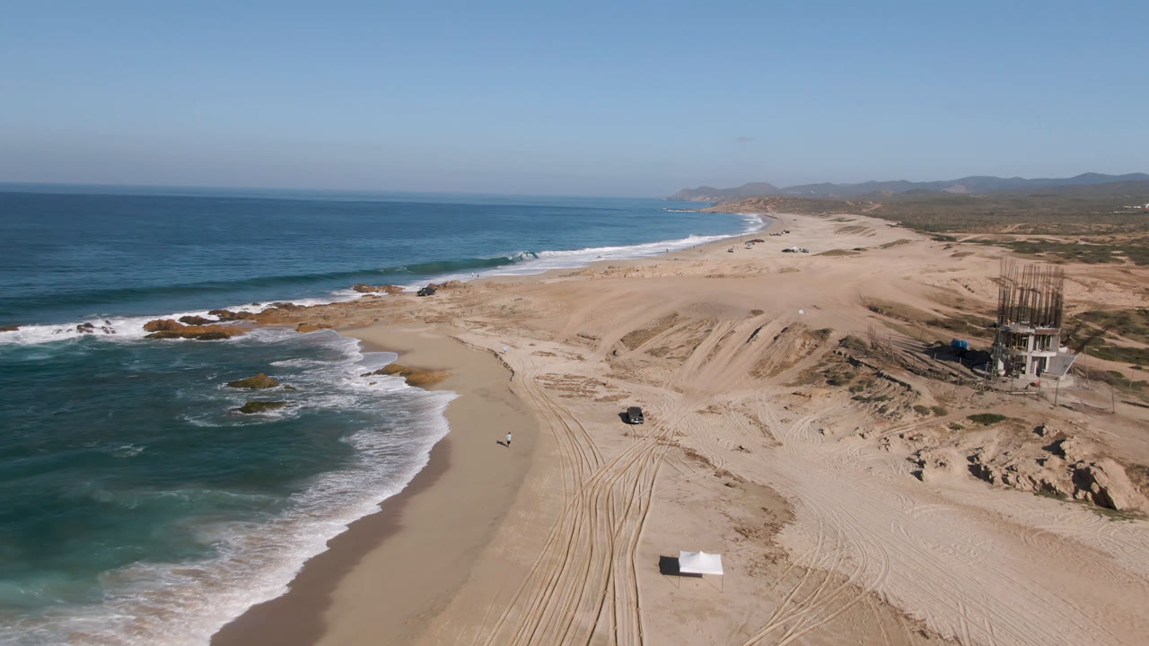 Drone-style aerial view in Migrino with blue water and white surf at left, a broad sandy beach with vehicle tracks, a dark truck below center, dunes to the right, and no visible people.