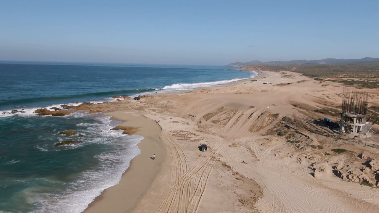High aerial view of a sandy shoreline with waves, dunes, tracks, and a truck.