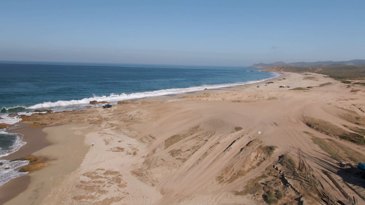 Opening aerial frame over Migrino, Baja California Sur shows broad tan sand, blue ocean surf, a small parked truck by the water, distant hills, and no visible people.