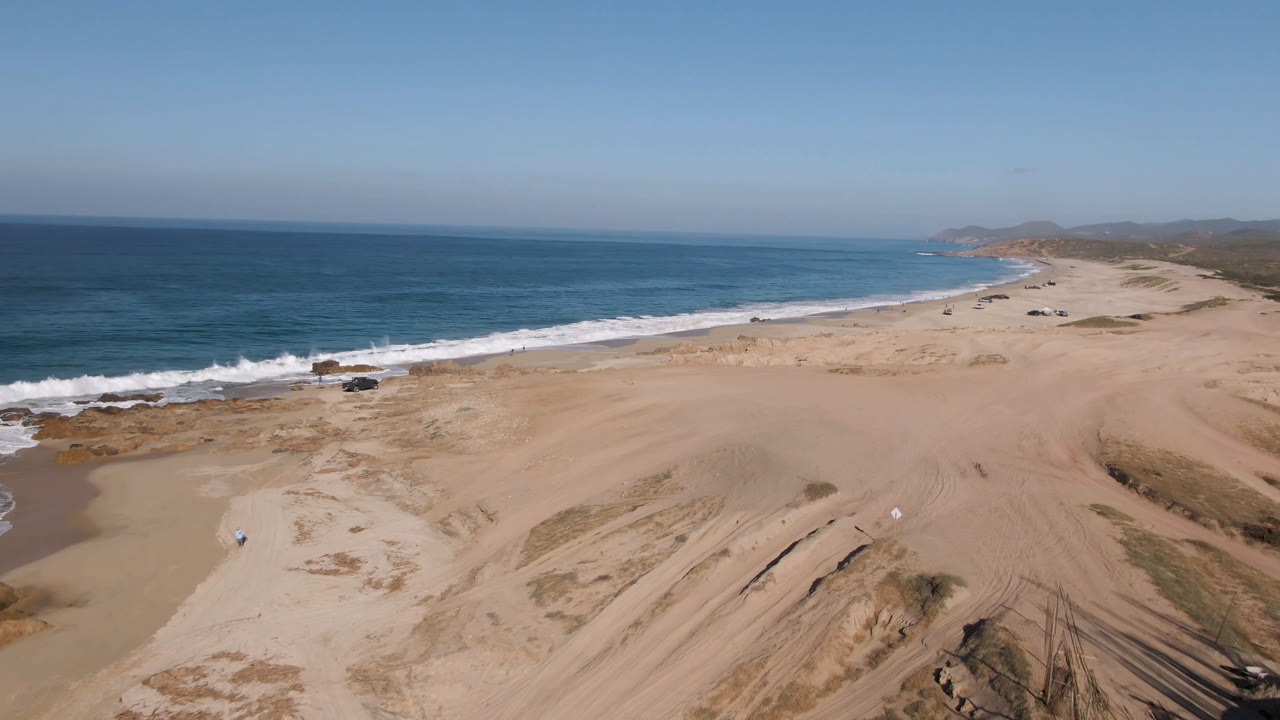 Second aerial frame in Migrino, Baja California Sur keeps the truck near the lower shoreline while sand ridges, vehicle tracks, blue water, and no visible people fill the view.