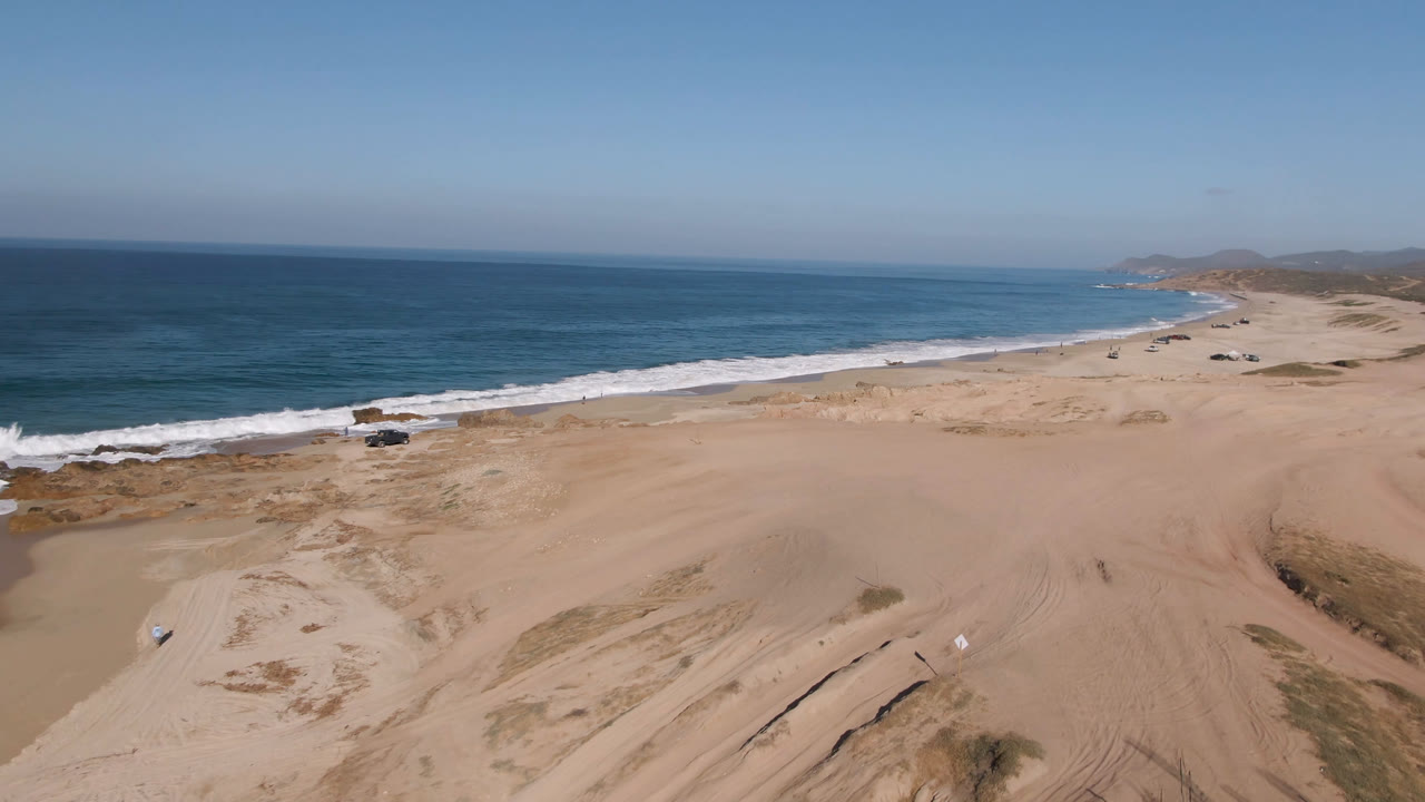 Wide aerial beach frame with surf, dunes, tracks, and a small truck by the water.