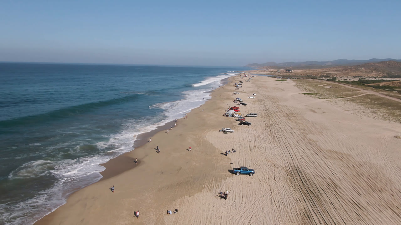 Aerial frame over Migrino, Baja California Sur, showing blue ocean surf on the left, a broad sandy beach on the right, parked vehicles near the waterline, and tiny distant figures.