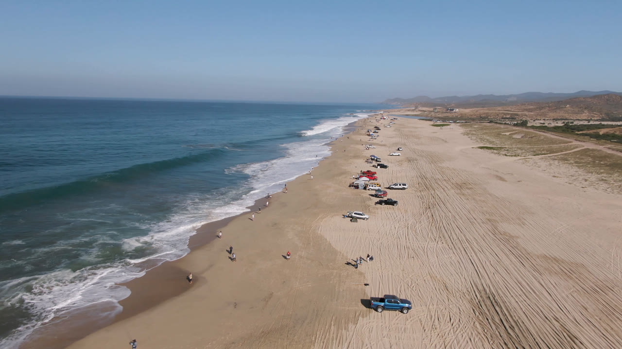 A second aerial frame over Migrino shows the shoreline stretching into the distance, vehicles and shade canopies grouped on the sand, and small surf breaking along the beach.