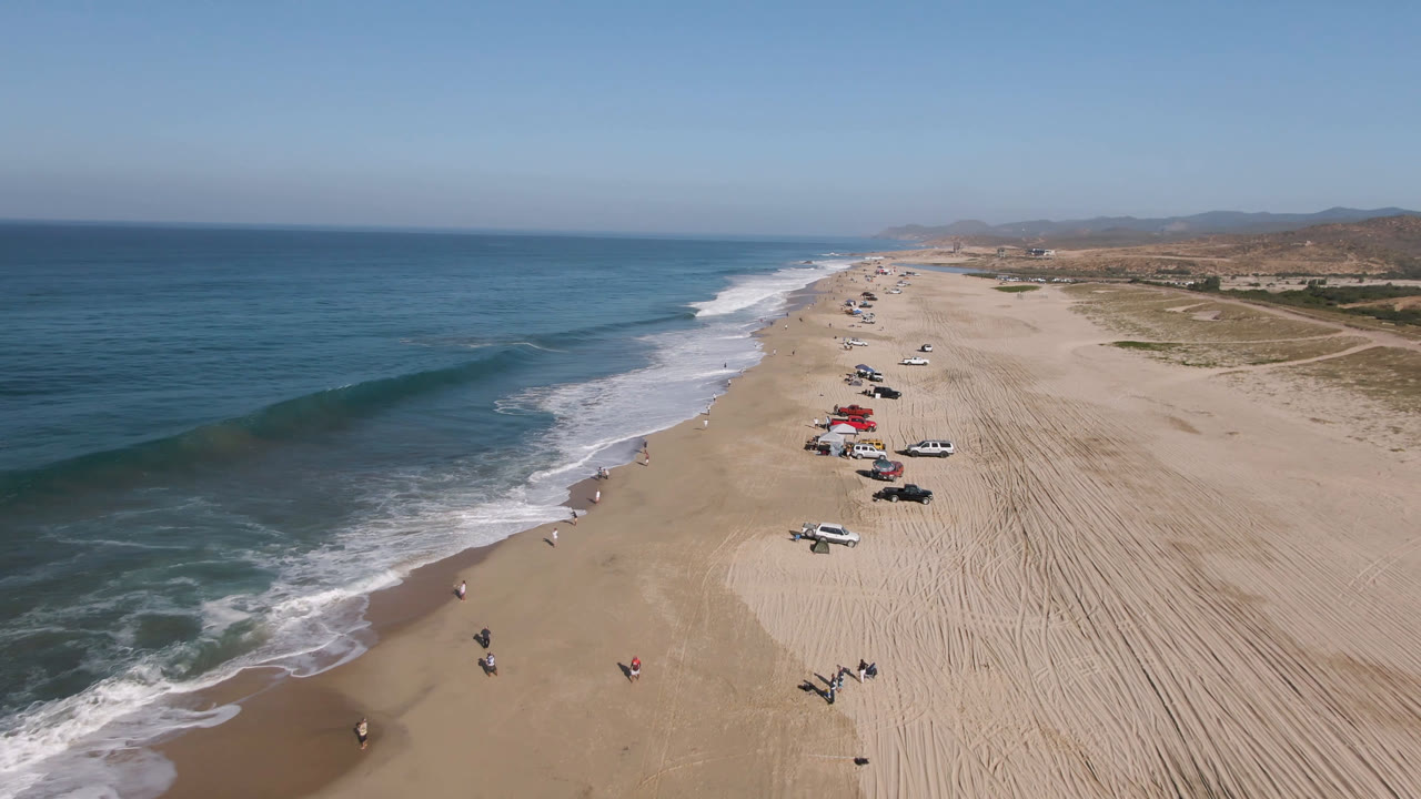 Wide aerial coastline with ocean waves, beach vehicles, and open sand.
