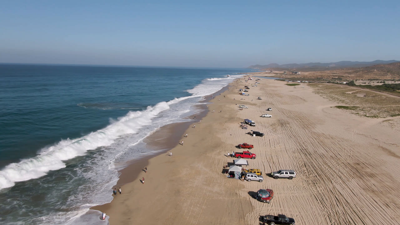Aerial view of surf, sandy beach, parked vehicles, and distant coastal hills.