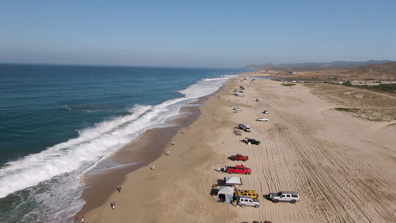 Drone view along Migrino beach as white waves reach the sand, with clusters of parked cars and trucks, a small canopy, patterned tire tracks, distant hills, and tiny distant beachgoers.