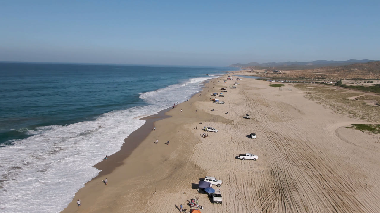 Aerial view over Migrino, Baja California Sur, with blue ocean and white surf beside a wide sandy beach where several cars are parked and tiny beachgoers are visible.