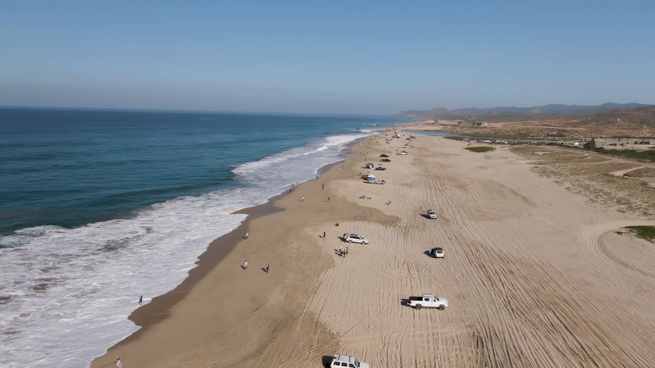 Drone-wide frame looking along the Migrino shoreline, with surf curling in from the left, tire-marked sand stretching inland, parked cars spaced along the beach, and small people visible near the waterline.
