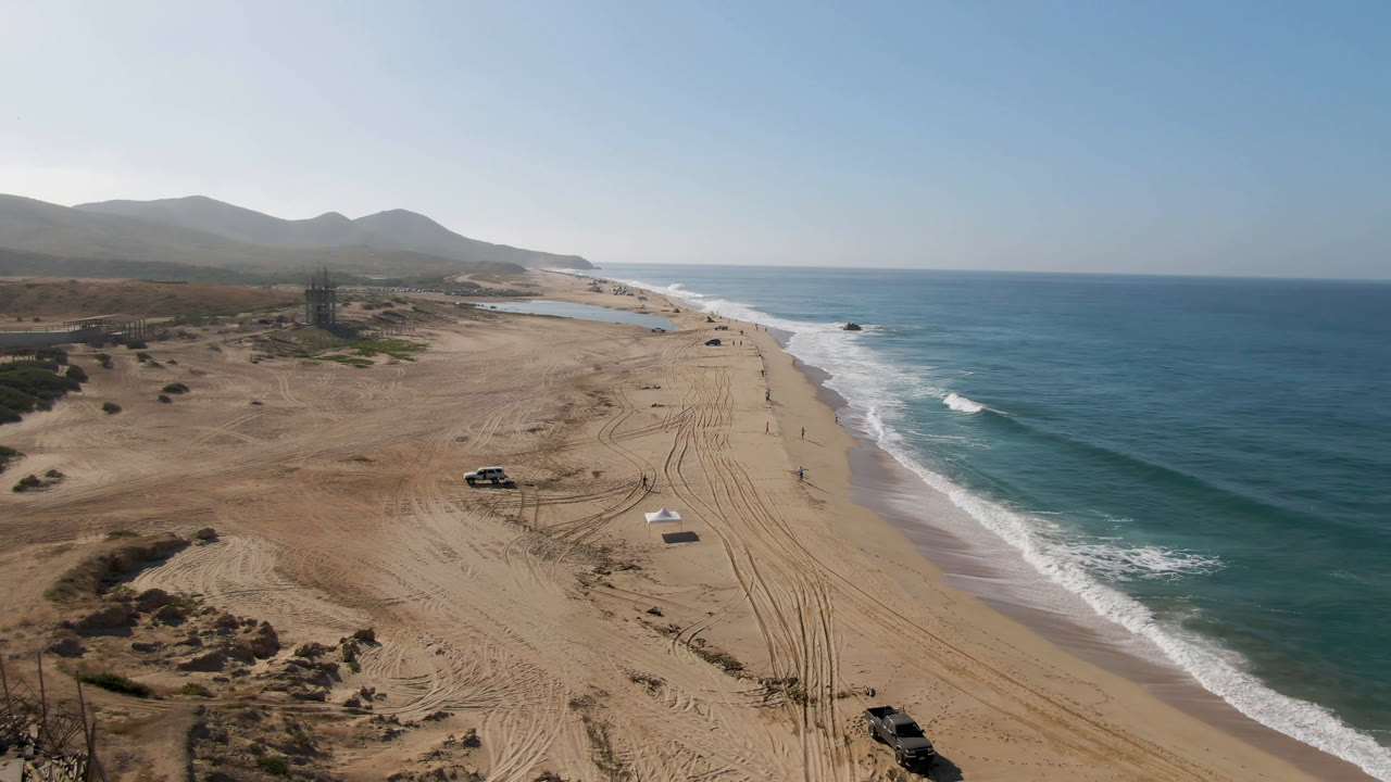 Aerial view over Migrino, Baja California Sur showing a sandy beach, a dark truck near the lower edge, parallel tire tracks, surf on the right, distant hills, and no visible people.