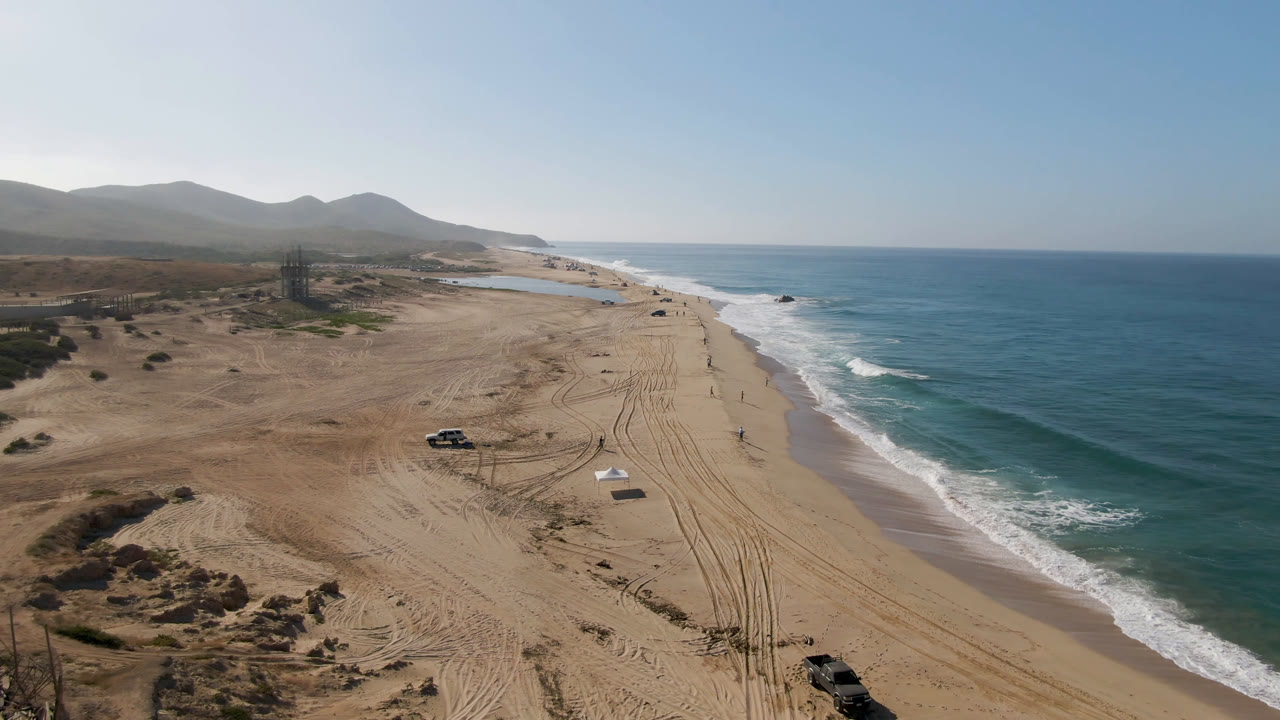 A high drone frame in Migrino, Baja California Sur shows the truck slightly farther along the sandy beach, with tire marks leading toward the surf and no visible people.