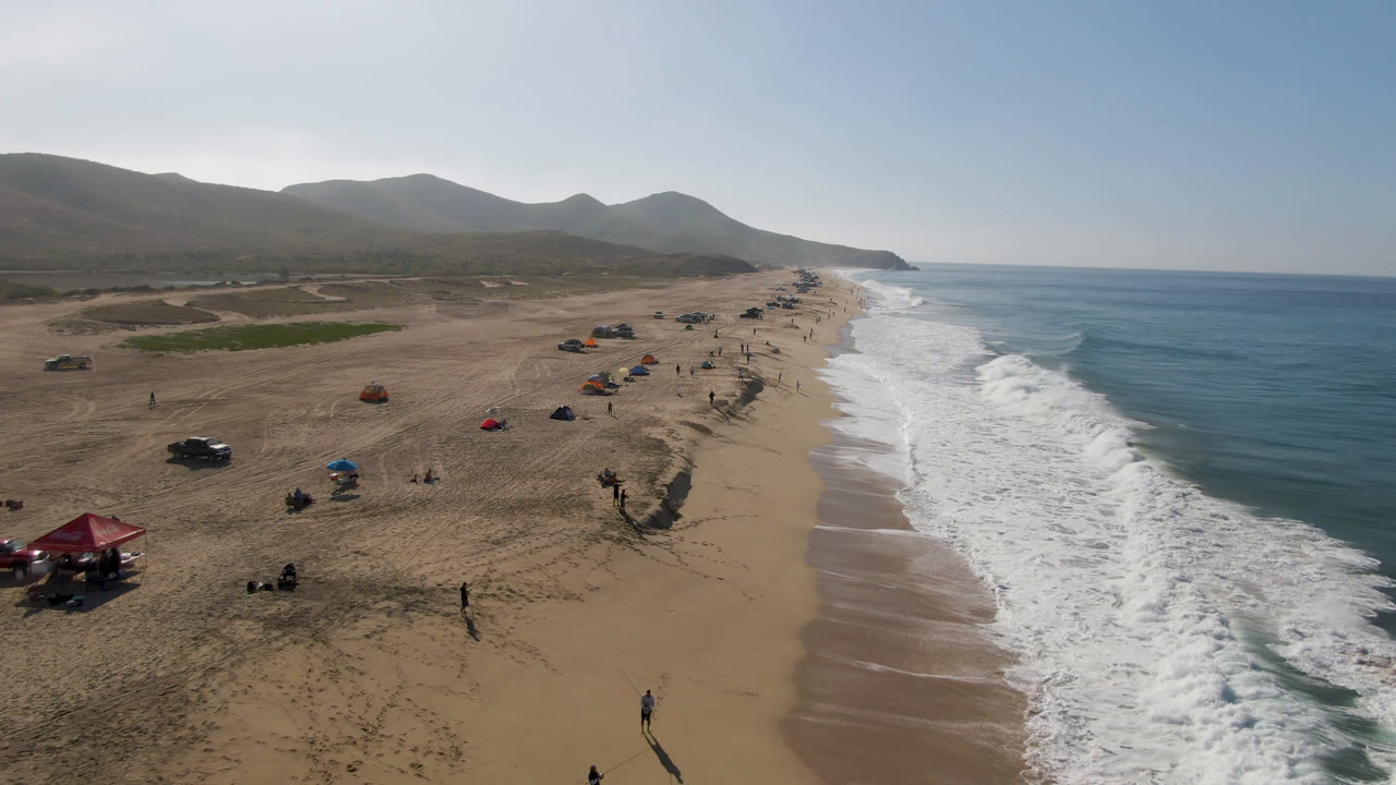 Aerial frame over Migrino, Baja California Sur, showing scattered beach umbrellas and people on wide sand beside white surf and blue ocean, with hazy hills behind the beach.