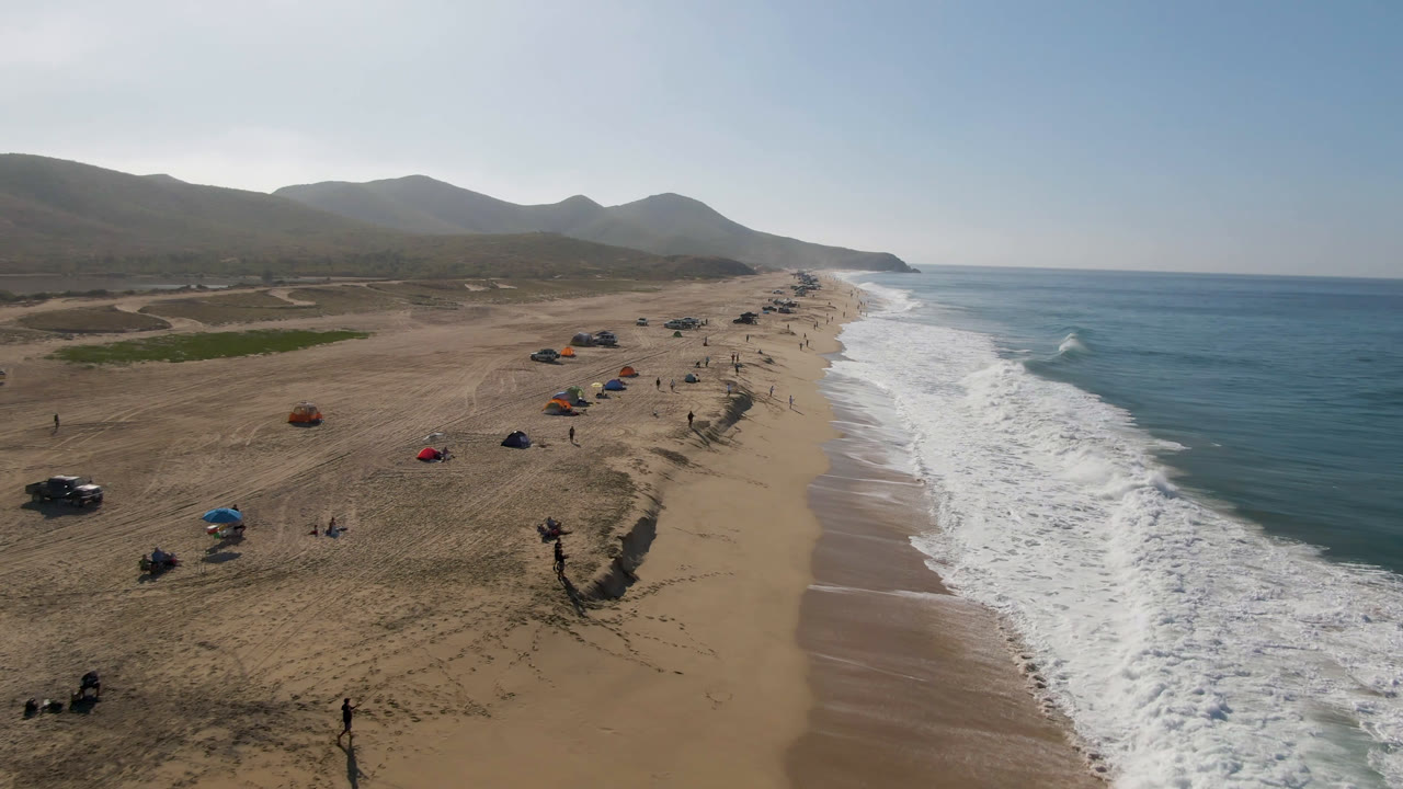Migrino aerial coastline view with beachgoers and small umbrellas running down the sand, parked vehicles set back from the water, and white waves breaking along the shore.