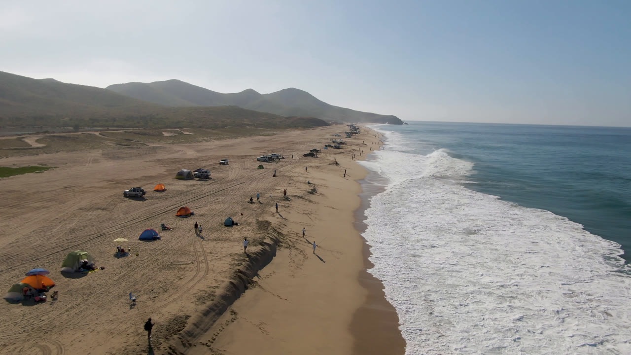 Aerial view of sandy beach, surf, umbrellas, vehicles, and distant mountains.