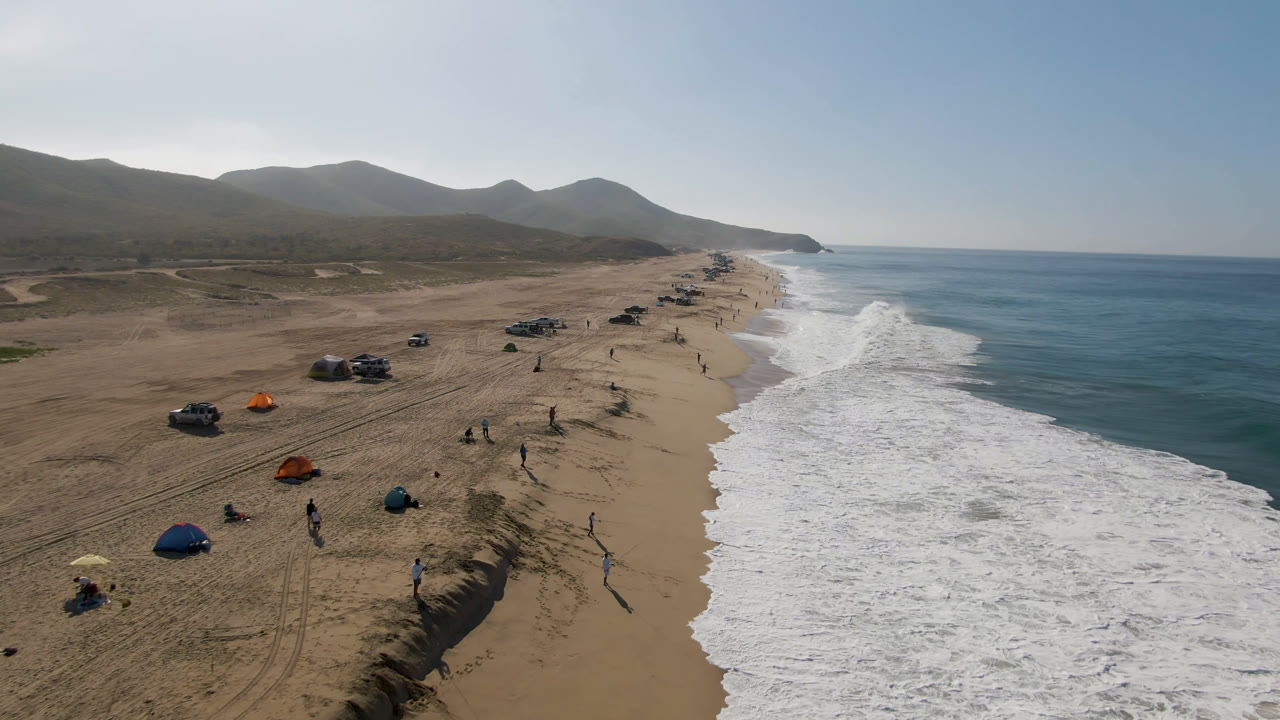 Aerial view in Migrino, Baja California Sur with the shoreline stretching into the distance, white waves breaking beside tan sand, and small beachgoers near umbrellas and vehicles.