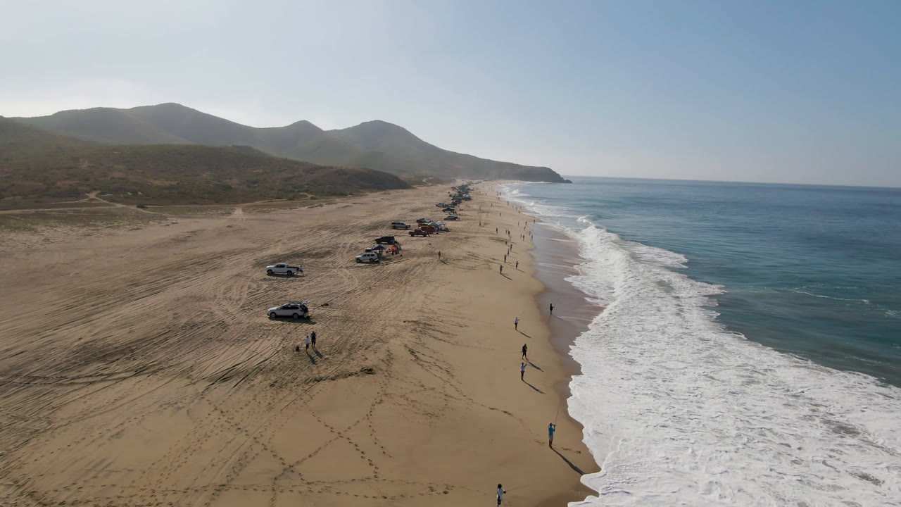Aerial view of sandy beach with parked cars, surf, ocean, and mountains.
