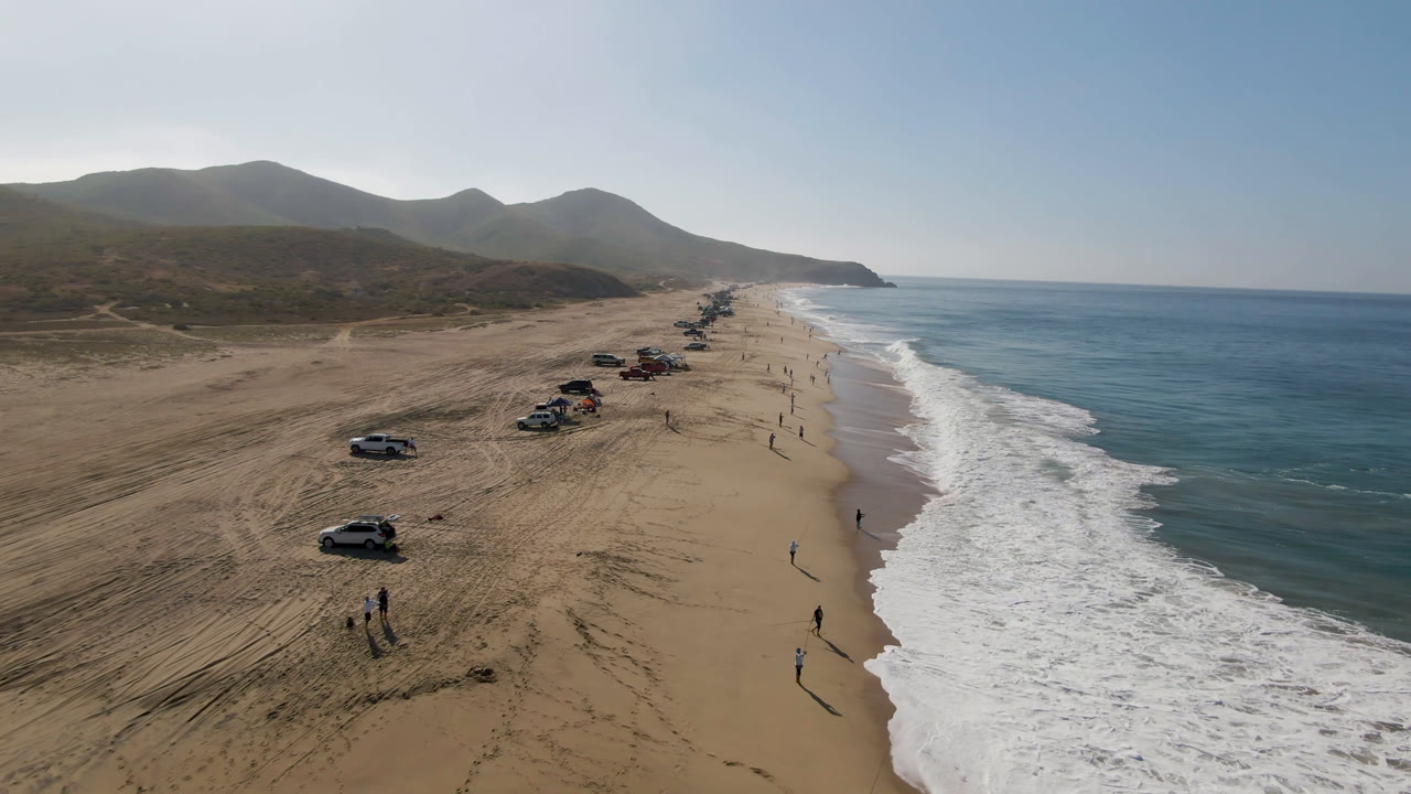 Drone view along Migrino beach with scattered vehicles on the sand, white waves breaking beside the shoreline, dry hills behind the beach, and small distant people visible on the wet sand.