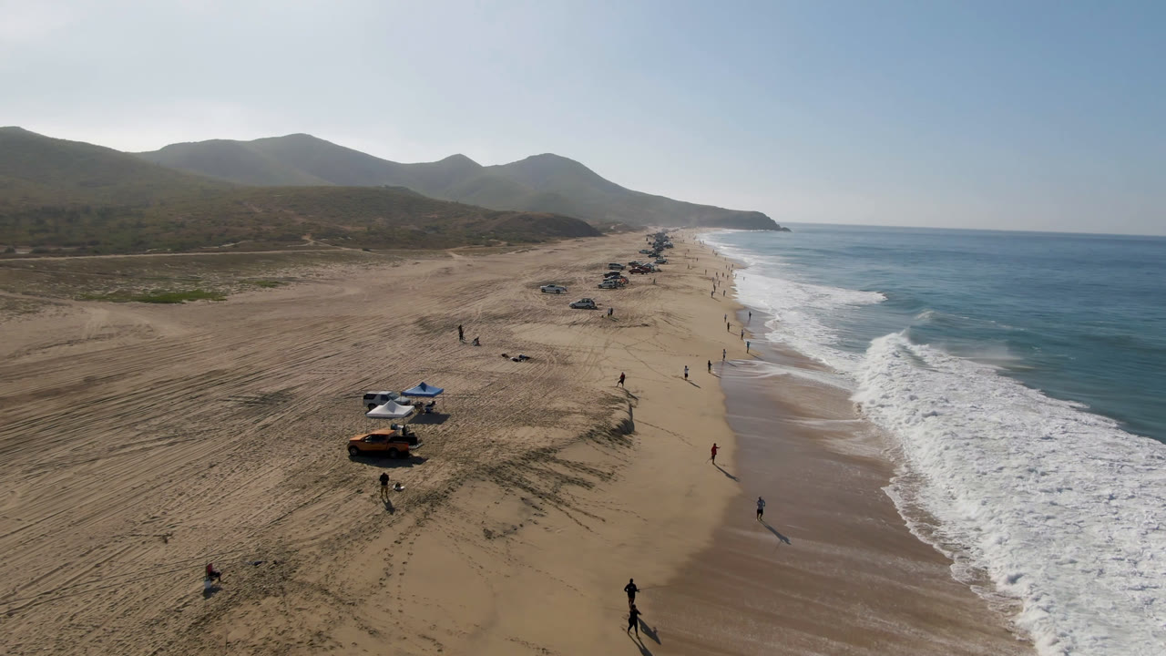 Aerial beach view with parked vehicles, surf, and hills.
