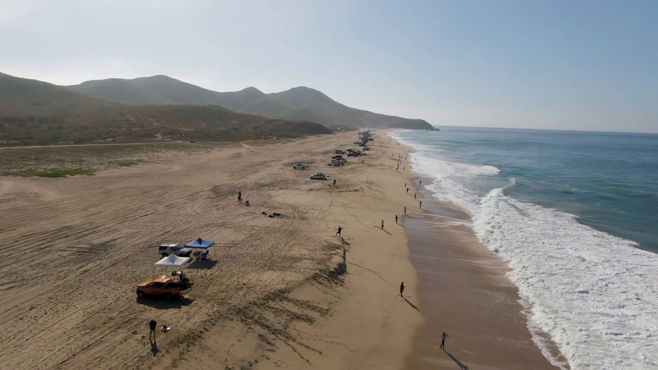 Drone view over Migrino beach with a line of parked vehicles on the sand, scattered distant people near the waterline, active white waves, and hills receding along the coast.