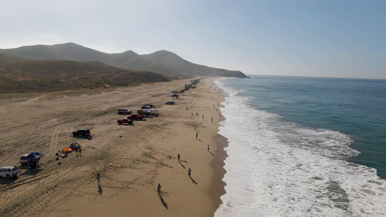 Aerial view of vehicles parked along a sandy beach beside ocean surf.
