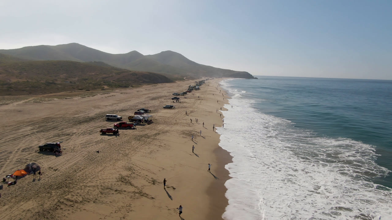 Aerial view in Migrino, Baja California Sur, Mexico, with trucks and SUVs grouped on the left side of the beach, foamy ocean water on the right, and low coastal hills in the background.
