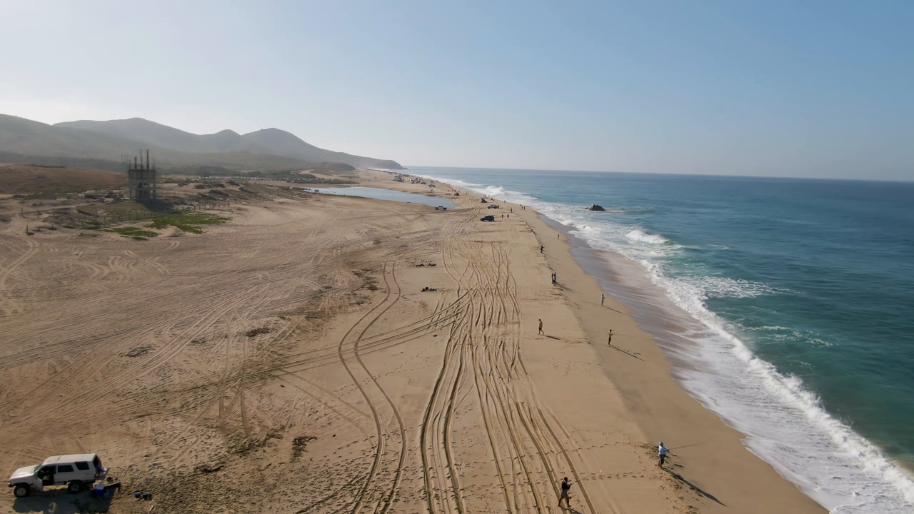 Wide aerial view of a sandy beach with tire tracks, surf, hills, and a vehicle at the lower left.