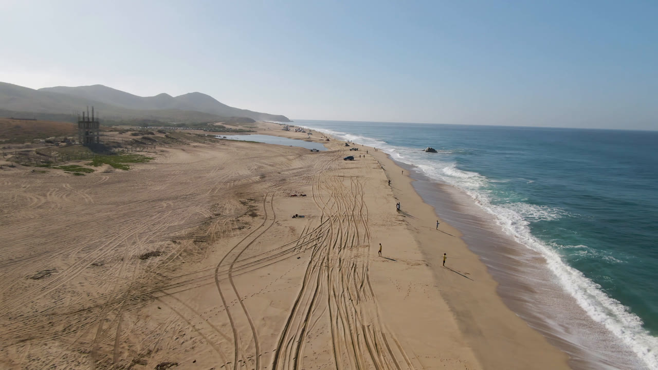 Drone view over Migrino beach with the shoreline running diagonally, parallel tire tracks across the sand, white surf along the right edge, hills and a partially built structure inland, and small distant people visible near the waterline.